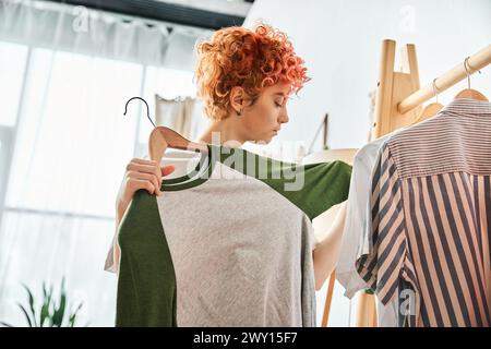 appealing red haired extravagant person picking up stylish clothes near rack at home, leisure time Stock Photo