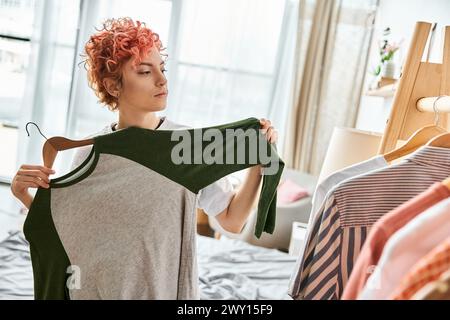 appealing red haired extravagant person picking up stylish clothes near rack at home, leisure time Stock Photo