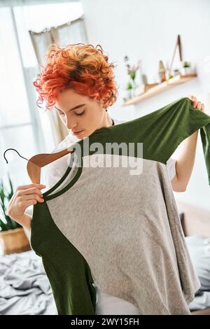 appealing red haired extravagant person picking up stylish clothes near rack at home, leisure time Stock Photo