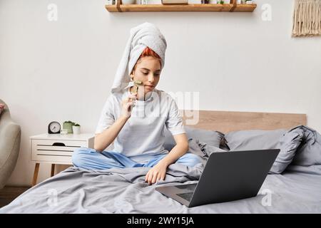 appealing red haired queer person in homewear using face roller while relaxing on bed with laptop Stock Photo