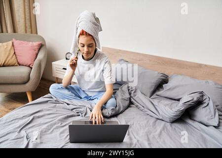 appealing red haired queer person in homewear using face roller while relaxing on bed with laptop Stock Photo