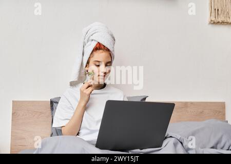 appealing red haired queer person in homewear using face roller while relaxing on bed with laptop Stock Photo