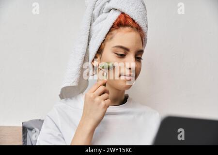 attractive red haired queer person in homewear using face roller while relaxing on bed with laptop Stock Photo