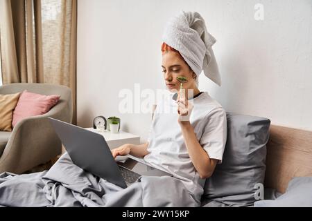 attractive red haired queer person in homewear using face roller while relaxing on bed with laptop Stock Photo