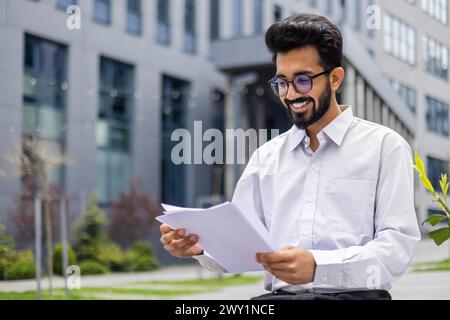 A smiling Indian man in a white shirt and glasses sits on a bench near the offices and reads the ,documents he is holding in his hands. Stock Photo