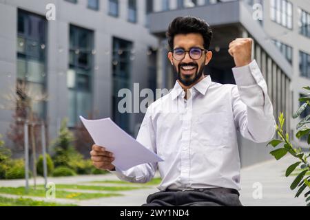 Portrait of a happy Indian male businessman smiling at the camera and happy reading a letter and documents he is holding in his hands showing a victory gesture. Stock Photo