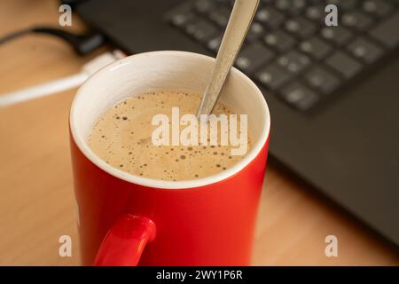 Red cup of coffee with crema and spoon in the cup standing besides a notebook. Just keyboard and some plugs can be seen from the notebook. Close up. Stock Photo