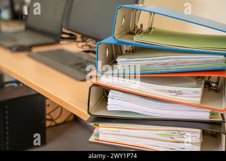 Pile of file folders stacked on desk, in front of computer and laptop of an office environment. Computers blurry. Not a paperless office. Stock Photo