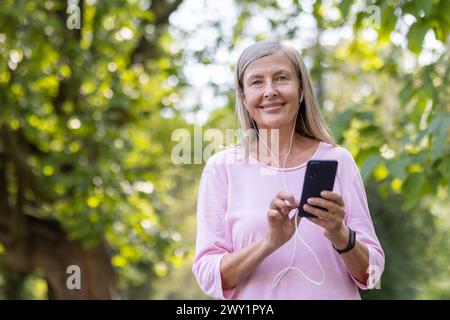 Close-up portrait of a smiling gray-haired sporty woman standing in the park wearing headphones, holding a phone and looking at the camera. Stock Photo