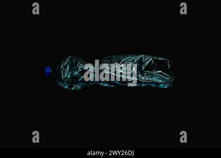 A crushed plastic bottle partially filled with water, lying horizontally on a black background. Studio light reflections highlight the edges of the cr Stock Photo