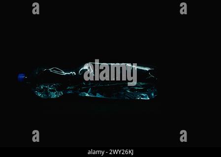 A crushed plastic bottle partially filled with water, lying horizontally on a black background. Studio light reflections highlight the edges of the cr Stock Photo