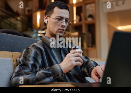 Portrait of focused young man wearing glasses and drinking coffee while working with laptop in office lounge or hotel lobby Stock Photo