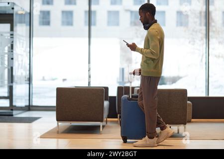 Side view full length portrait of African American man with luggage walking across hotel lobby and using smartphone calling taxi app Stock Photo