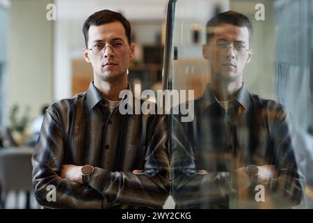 Waist up portrait of confident Asian man wearing glasses leaning on glass wall in office with reflection and looking at camera Stock Photo