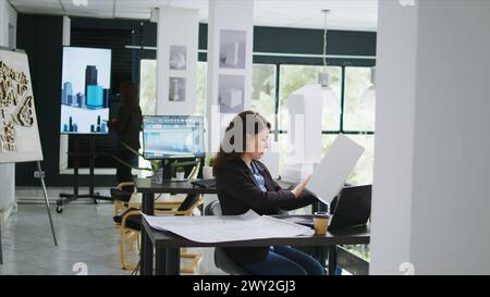 Architectural technician examines 3d building model in creative agency coworking space, taking notes of design lines and measurements. Woman measuring maquette scale and elements. Tripod shot. Stock Photo
