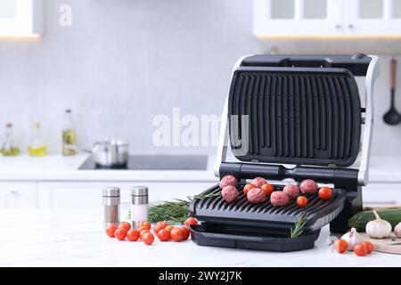 Electric grill with meatballs, tomatoes, spices and vegetables on white marble table in kitchen. Space for text Stock Photo