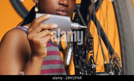 BIPOC technician using specialized lubricant to repair squeaky bicycle chain, orange studio background. Specialist applying oil on noisy bike parts during maintenance process, close up shot Stock Photo