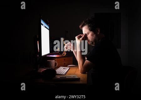 Tired and stressed office worker illuminated by the glow of their computer screen in a dark room at night while working late. Burn out. Stock Photo