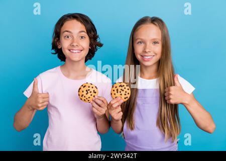 Photo of excited cool friends wear casual outfits showing thumbs up eating tasty cookies isolated blue color background Stock Photo