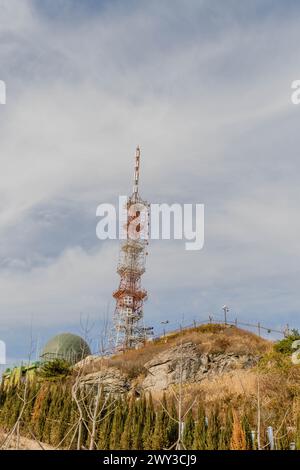 A tall communication tower perched on a hill with surrounding greenery under a cloudy sky, in South Korea Stock Photo