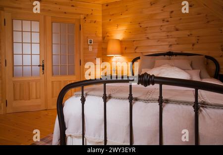 Queen size bed with white bedspread, throw and pillows, tubular cast iron headboard and footboard in master bedroom with pine wood plank walls Stock Photo