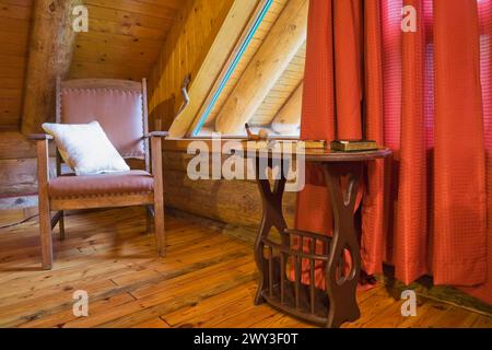Brown wooden leather armchair with white cushion and side table in master bedroom with red curtains and wood plank floor on upstairs floor inside Stock Photo