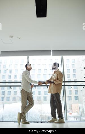 Vertical wide angle shot of two young male colleagues shaking hands standing against floor to ceiling window in office Stock Photo