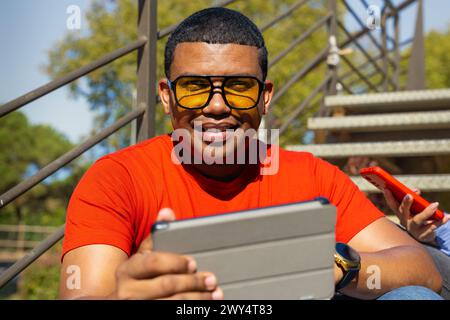 Glamorous smiling young man, sitting using a app on a digital tablet outdoors. Stock Photo