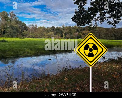 Radioactive pollution. Yellow warning sign with hazard symbol near river outdoors Stock Photo