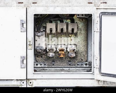 A close-up view of an old and corroded electrical fuse box mounted on a deteriorating exterior wall. The box is open, exposing fuses and connectors, w Stock Photo