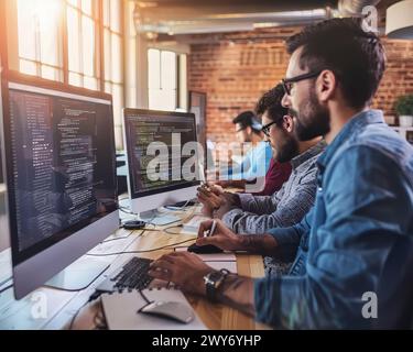A group of men are working on computers in a room. They are all wearing glasses and are focused on their work. The room has a brick wall and a window Stock Photo