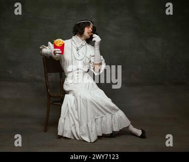 Portrait of man dressed old-fashioned dress and eating French fries from modern restaurant against vintage studio background. Stock Photo