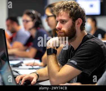 A man with a beard is sitting at a desk with a laptop. He is wearing a watch and a black shirt. The man is focused on his work, possibly typing or bro Stock Photo