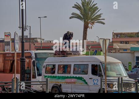 An image of a Moroccan citizen on top of a van loaded with people on a street in Marrakech tying bags and quilts on a street full of traffic Stock Photo