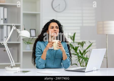A woman in a blue shirt enjoys a coffee break at her clean, white office space with a laptop and indoor plants. Stock Photo