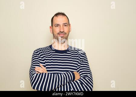 Bearded Hispanic man in his 40s wearing a striped sweater keeping his arms crossed and smiling while isolated on beige background. Stock Photo