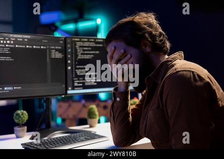 Troubled man facepalming himself while doing software quality assurance, finding major errors in source code. Depressed developer upset after inspecting coding on desktop PC and seeing many issues Stock Photo
