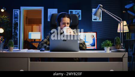 Caucasian man sitting on chair at home office desk in neon illuminated apartment, working and listening music. Teleworker wearing headphones in RGB lit living room, doing his job remotely Stock Photo