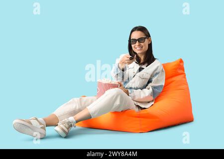 Beautiful young happy woman with bucket of popcorn watching TV on beanbag chair against blue background Stock Photo