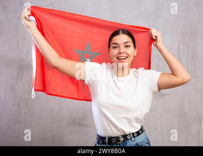 Happy young woman posing and smiling, standing with flag of Morocco against gray wall Stock Photo