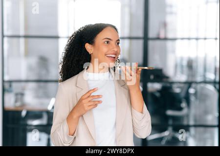 Happy beautiful brazilian or latino elegant curly haired woman, secretary, company ceo, stand in a modern office space, using smart phone, recording audio message, speaks on speakerphone, smiles Stock Photo
