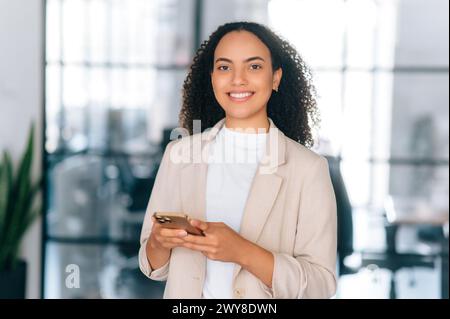 Cheerful pretty brazilian or hispanic business woman, company employee, secretary, stand in modern office space, using mobile phone for chatting with client or partner, looks at camera, smiles Stock Photo