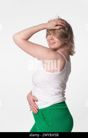 Portrait of happy plump woman is smiling and looking over her shoulder at camera, posing with one hand raised to head and hand on stomach. She wearing Stock Photo