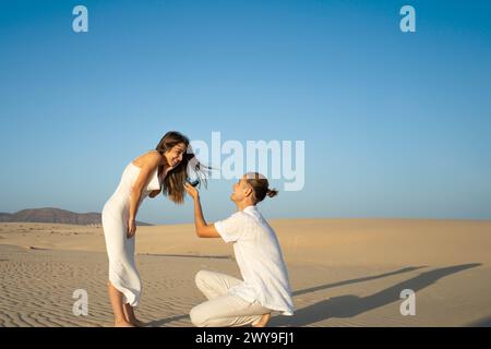 Joyful woman accepts a proposal on a sandy beach at sunset, as her partner kneels Stock Photo