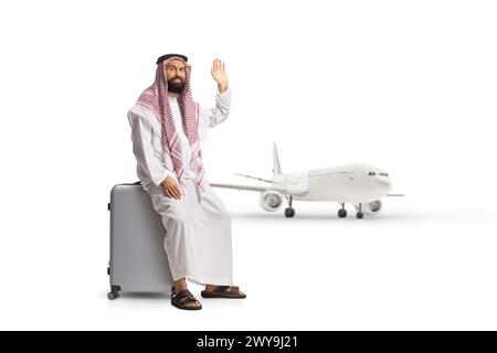 Saudi arab man sitting on a suitcase near an airplane and waving isolated on white background Stock Photo