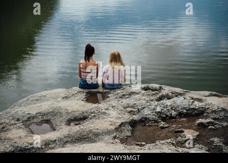 Wide rear view of two teen girls sitting on rocks near a lake. Stock Photo