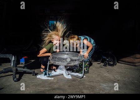 Two siblings lean over fan with hair blowing in garage Stock Photo