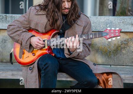 long-haired European guy plays electric guitar sitting on bench Stock Photo