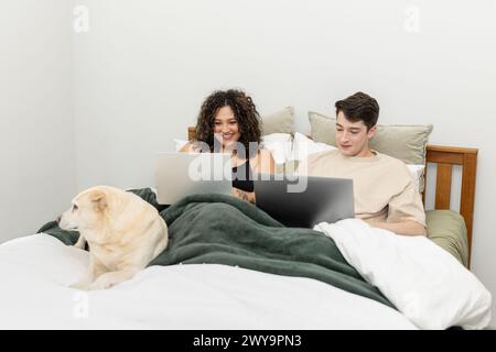 Smiling couple works side by side on laptops in bed with their dog Stock Photo