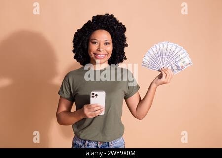 Portrait of positive person with perming coiffure hold smartphone bunch of dollars in arm get salary isolated on pastel color background Stock Photo